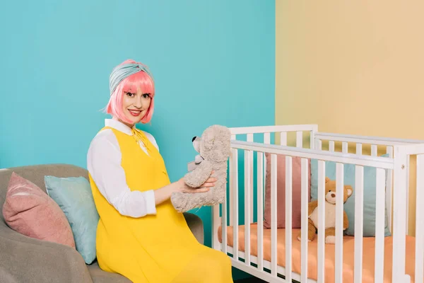 Happy retro styled pregnant pin up woman with pink hair sitting with teddy bear near baby cot — Stock Photo