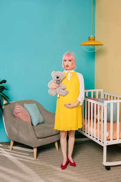 Style rétro goupille enceinte femme avec des cheveux roses debout avec ours en peluche dans la chambre d'enfant — Photo de stock