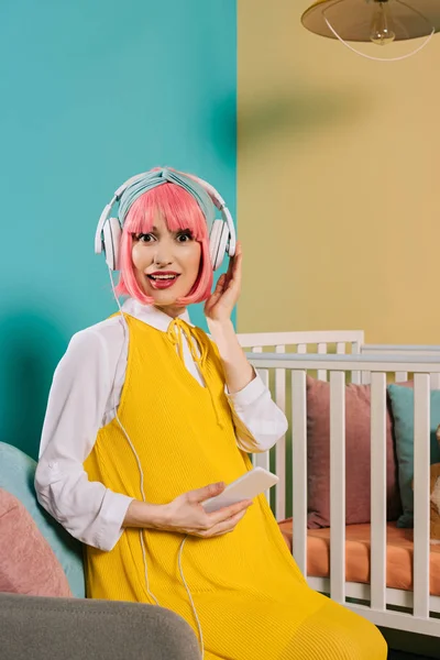 Здивована вагітна жінка з рожевим волоссям слухає музику біля дитячого ліжечка в дитячій кімнаті — стокове фото
