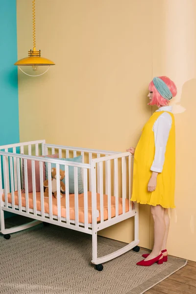 Vue latérale de style rétro femme enceinte pin up avec des cheveux roses debout près de lit bébé dans la chambre d'enfant — Photo de stock