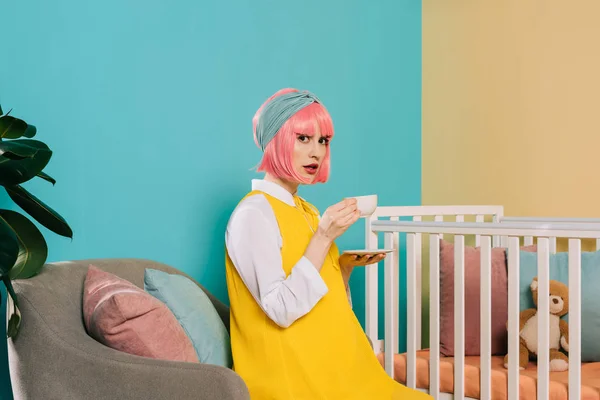 Retro styled pregnant pin up woman with pink hair sitting near baby cot with cup of tea and looking at camera — Stock Photo