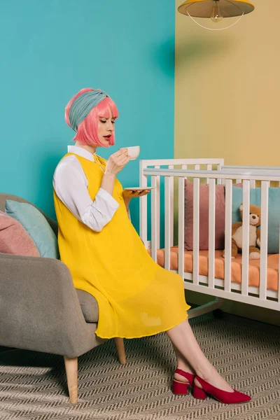 Style rétro pin up enceinte femme aux cheveux roses boire du thé près de lit bébé dans la chambre d'enfant — Photo de stock