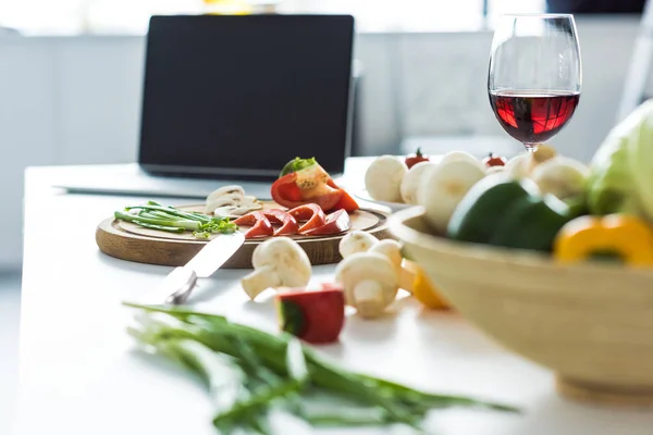 Laptop, copo de vinho tinto e legumes na mesa da cozinha — Fotografia de Stock