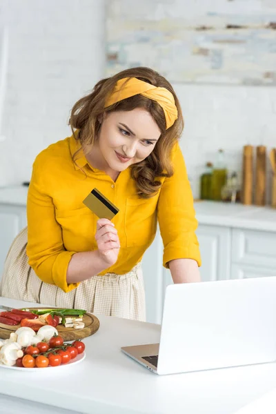 Sonriente hermosa mujer sosteniendo tarjeta de crédito y mirando a la computadora portátil en la cocina - foto de stock