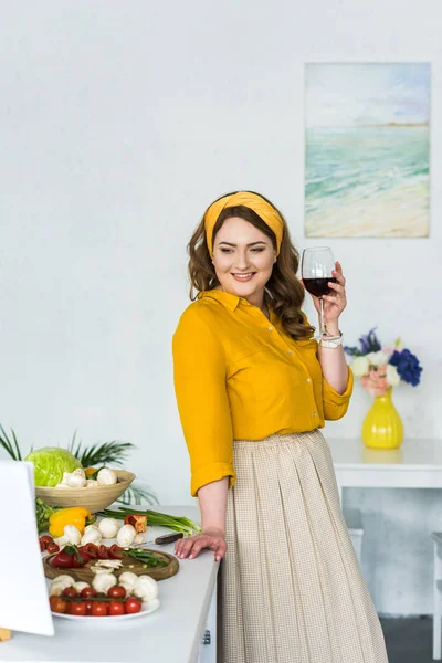 Belle femme tenant un verre de vin et regardant les légumes dans la cuisine — Photo de stock