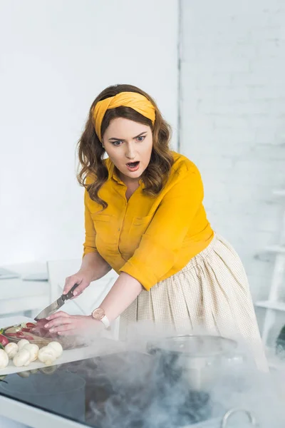 Потрясенная красивая женщина смотрит на горящую сковороду на электрической плите на кухне — стоковое фото