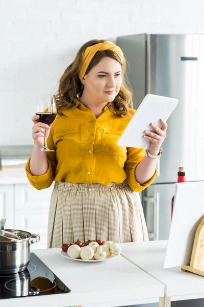 Красивая женщина с бокалом красного вина и с помощью планшета на кухне — стоковое фото