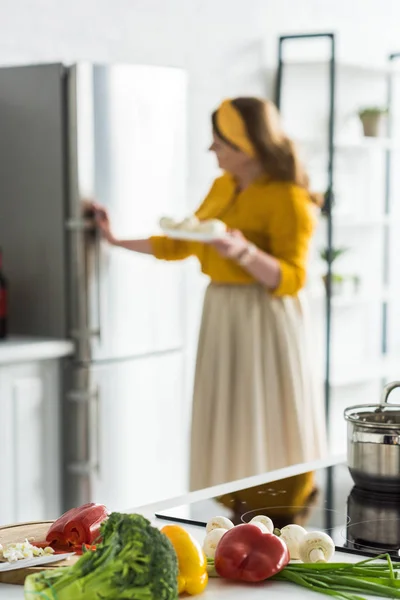 Woman taking plate from fridge with vegetables on foreground in kitchen — Stock Photo