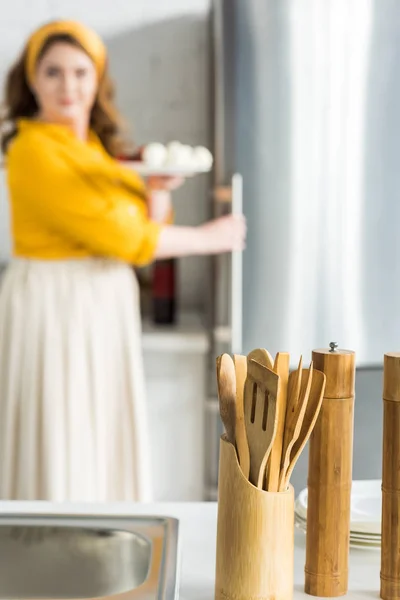 Woman taking plate from fridge with wooden spatulas on foreground in kitchen — Stock Photo