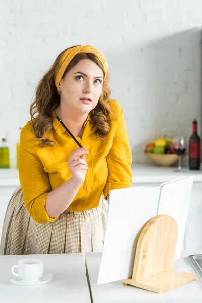 Nachdenkliche schöne Frau, die in der Küche in der Nähe von Rezeptbüchern nachschlägt — Stockfoto