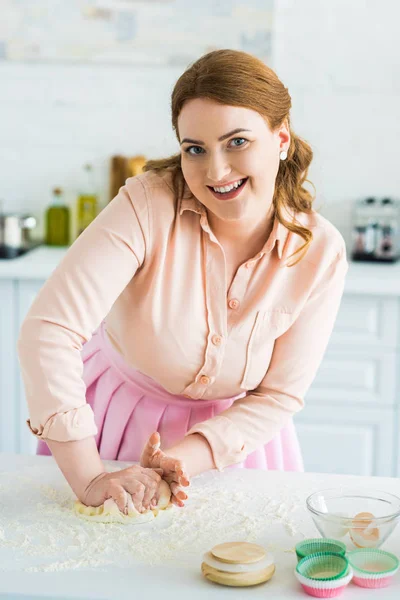 Sonriente hermosa mujer amasando masa en la cocina y mirando a la cámara - foto de stock