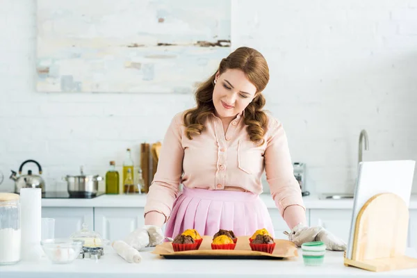 Sorrindo bela mulher olhando para bandeja com muffins na cozinha — Fotografia de Stock