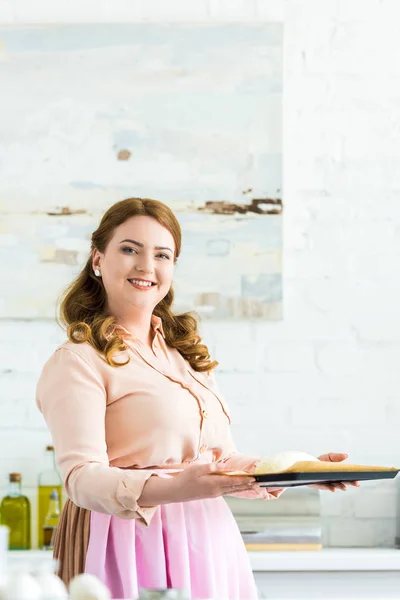 Frau hält Blech mit Teig zum Brotbacken und blickt in Küche in die Kamera — Stockfoto