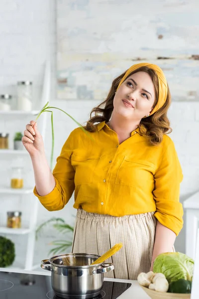 Pensivo bela mulher segurando cebola verde na cozinha — Fotografia de Stock