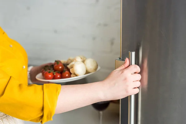 Immagine ritagliata della donna che apre il frigorifero e tiene il piatto con funghi e pomodori in cucina — Foto stock
