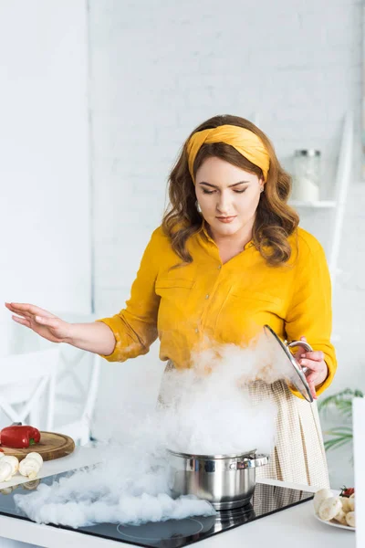 Bela mulher segurando tampa e olhando para o vapor da panela no fogão elétrico na cozinha — Fotografia de Stock