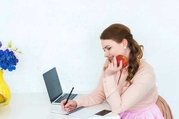 Belle femme écrit quelque chose pour ordinateur portable et tenant pomme à la cuisine — Photo de stock