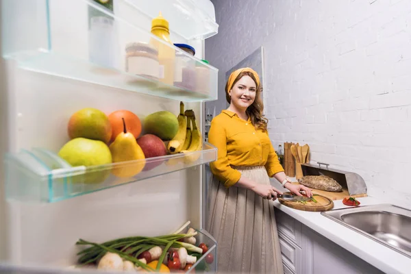 Bela mulher cortando legumes e olhando para a câmera na cozinha — Fotografia de Stock