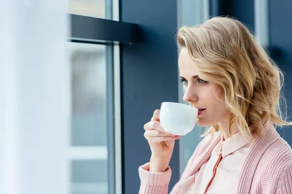 Vista laterale di donna premurosa bere caffè e guardando fuori dalla finestra — Foto stock