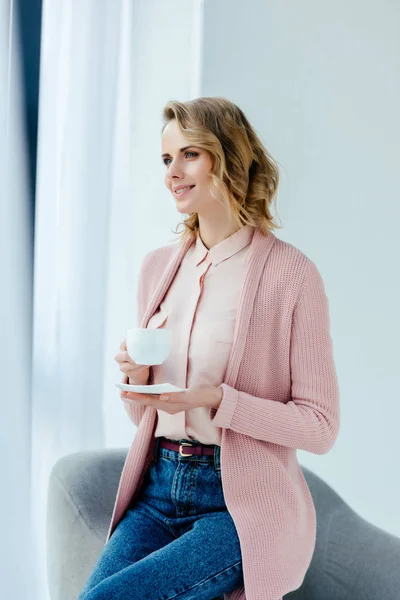 Retrato de mulher sorridente com xícara de café sentado na poltrona e olhando para longe — Fotografia de Stock