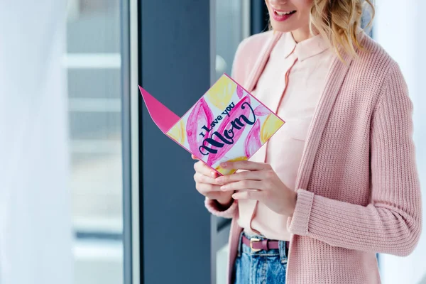 Partial view of smiling woman reading i love you mom postcard in hands, mothers day holiday concept — Stock Photo