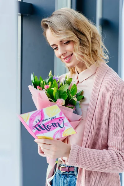 Sonriente mujer con ramo de flores leyendo te amo mamá postal, madre día concepto - foto de stock