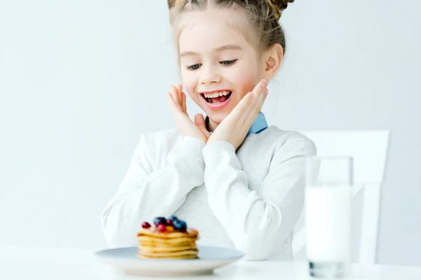 Fuoco selettivo del bambino eccitato guardando frittelle fatte in casa con bacche e miele sul tavolo — Foto stock