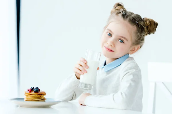 Portrait d'un enfant souriant avec un verre de lait à la main et des crêpes maison avec du miel et des baies sur la table — Photo de stock