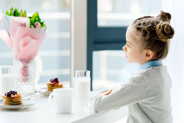 Vista lateral del niño sentado en la mesa con ramo de flores, vaso de leche y panqueques caseros - foto de stock