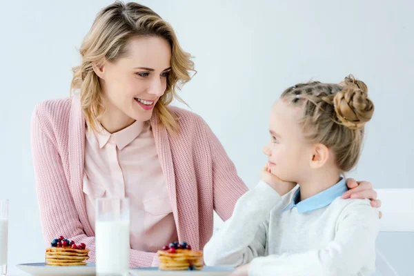 Retrato de mãe e filha sentadas à mesa com panquecas e copo de leite para o café da manhã — Fotografia de Stock