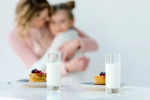 Enfoque selectivo de vasos de leche y panqueques en la mesa con abrazos familiares detrás - foto de stock