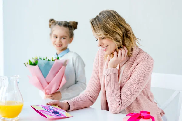 Selektiver Fokus der glücklichen Mutter auf Grußkarte und Tochter mit Blumenstrauß in der Nähe, Muttertagskonzept — Stockfoto