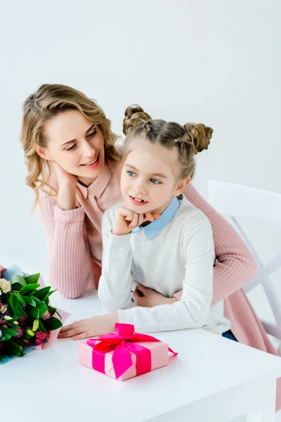 Madre abbraccio figlia a tavola con confezione regalo e bouquet, felice concetto di mamma giorno — Foto stock