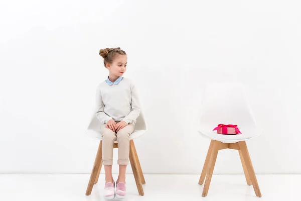 Adorable child sitting on chair and looking at present on another chair on white — Stock Photo