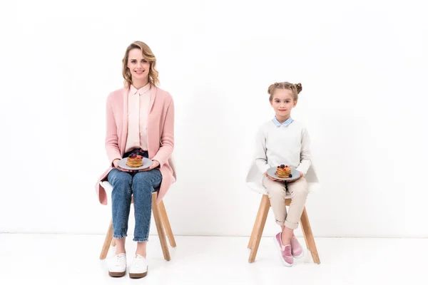 Madre e figlia sedute su sedie e che tengono piatti con frittelle su bianco — Foto stock
