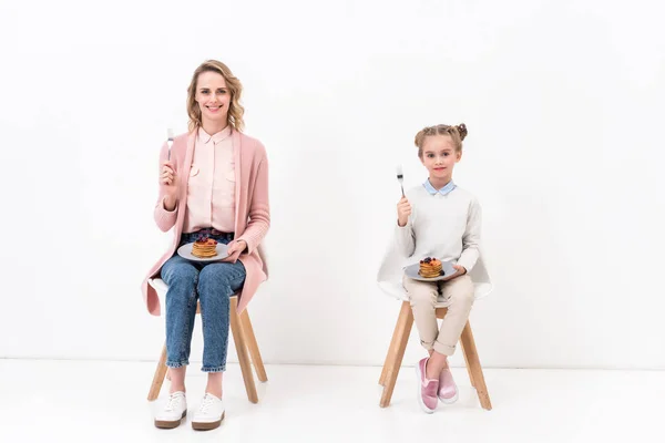 Mother and daughter sitting on chairs and holding forks and plates with pancakes on white — Stock Photo