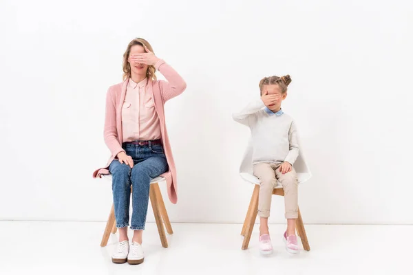 Mother and daughter sitting on chairs and covering eyes with hands on white — Stock Photo