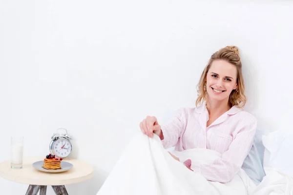 Attraktive Frau, die morgens im Bett liegt und in die Kamera schaut, Pfannkuchen auf dem Tisch neben dem Bett — Stockfoto