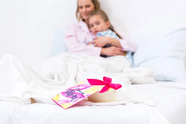 Mother and daughter lying on bed with present and postcard on foreground, happy mothers day concept — Stock Photo