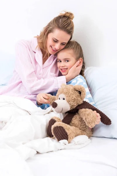 Mother hugging daughter with teddy bear on bed at home — Stock Photo