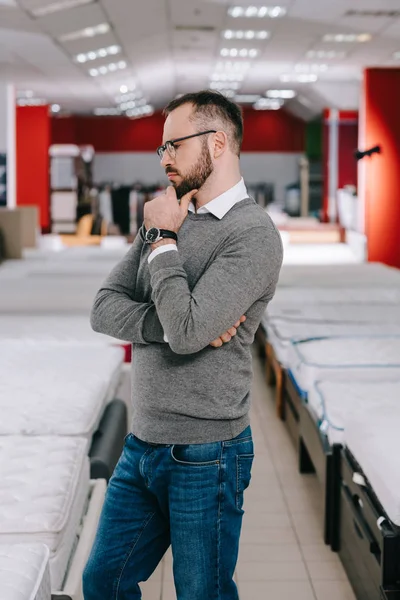 Seitenansicht eines nachdenklichen männlichen Kunden in Brille im Möbelhaus mit angeordneten Matratzen — Stockfoto