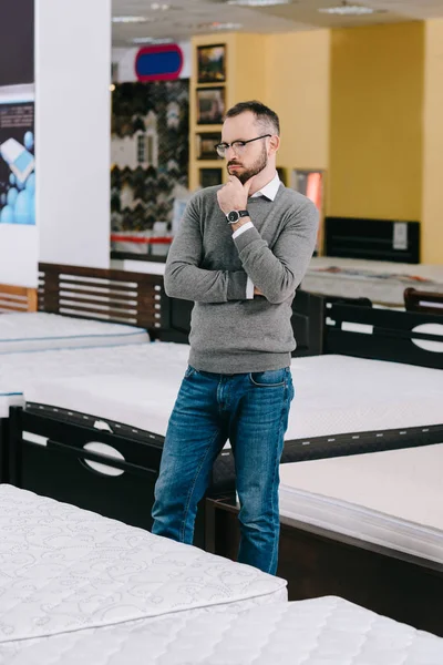 Cliente masculino pensativo en las gafas que eligen el colchón ortopédico en la tienda de muebles - foto de stock