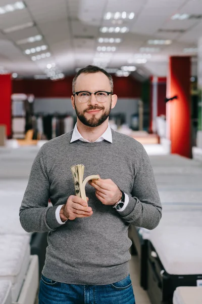 Portrait d'un homme souriant dans des lunettes avec des billets en dollars dans un magasin de meubles — Photo de stock