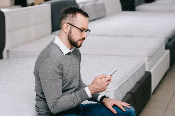 Side view of male shopper with smartphone sitting on mattress in furniture shop — Stock Photo