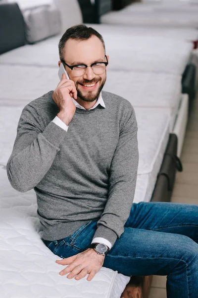 Ein lächelnder Mann, der auf einer Matratze im Möbelhaus sitzt und mit dem Smartphone spricht — Stockfoto