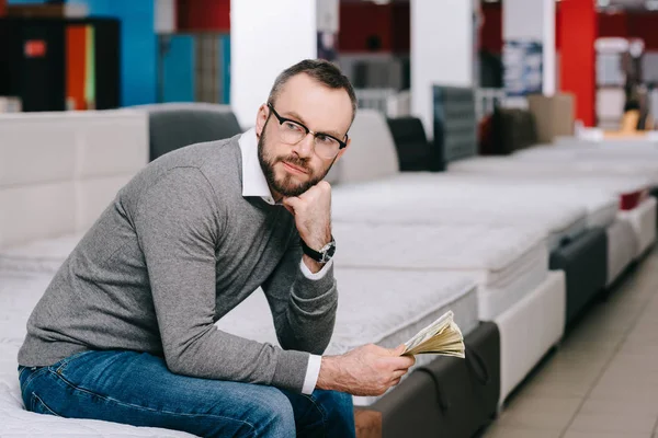 Side view of pensive male shopper with dollar banknotes sitting on mattress in furniture store — Stock Photo