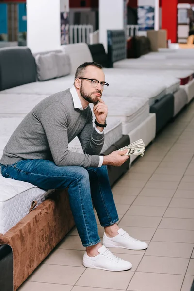 Side view of pensive male shopper with dollar banknotes sitting on mattress in furniture store — Stock Photo