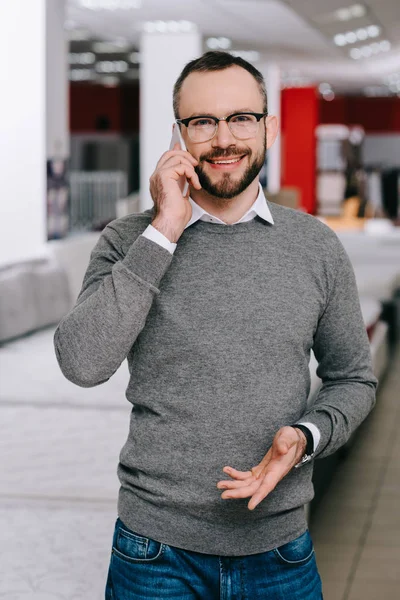 Portrait of man in eyeglasses talking on smartphone in furniture store with arranged mattresses — Stock Photo