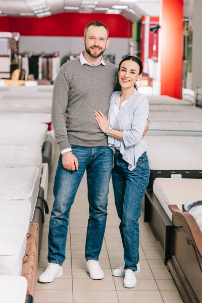 Smiling couple looking at camera in furniture store with arranged mattresses — Stock Photo