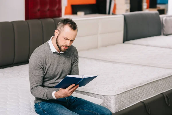 Konzentrierter Mann liest Notizen in Notizbuch im Möbelgeschäft mit arrangierten orthopädischen Matratzen — Stockfoto